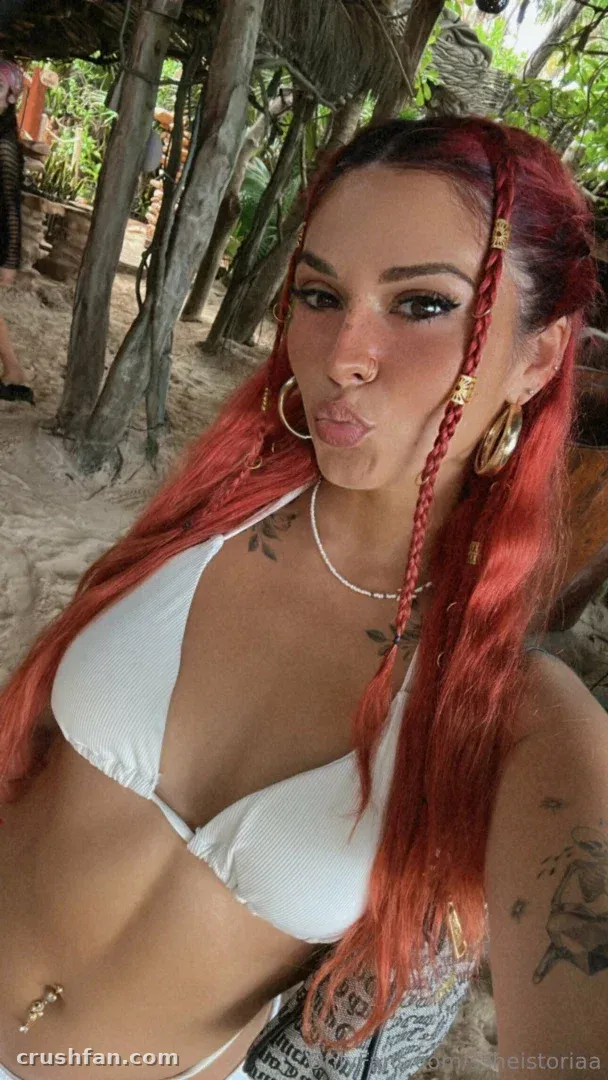 Toria Lorraine em um biquíni branco com tranças ruivas e brincos de argola dourados ao ar livre.