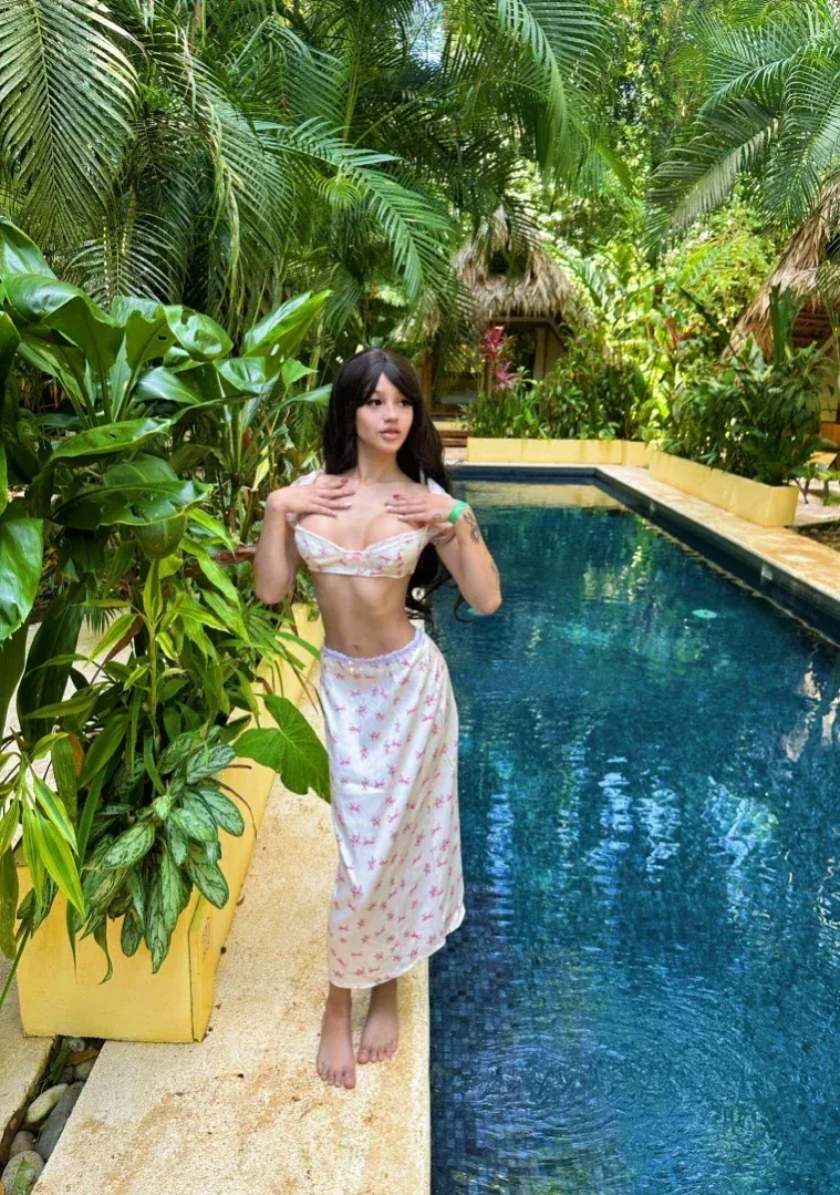 Paygeblog se tient pieds nus près d'une piscine dans un haut floral et une jupe longue au milieu de plantes tropicales.