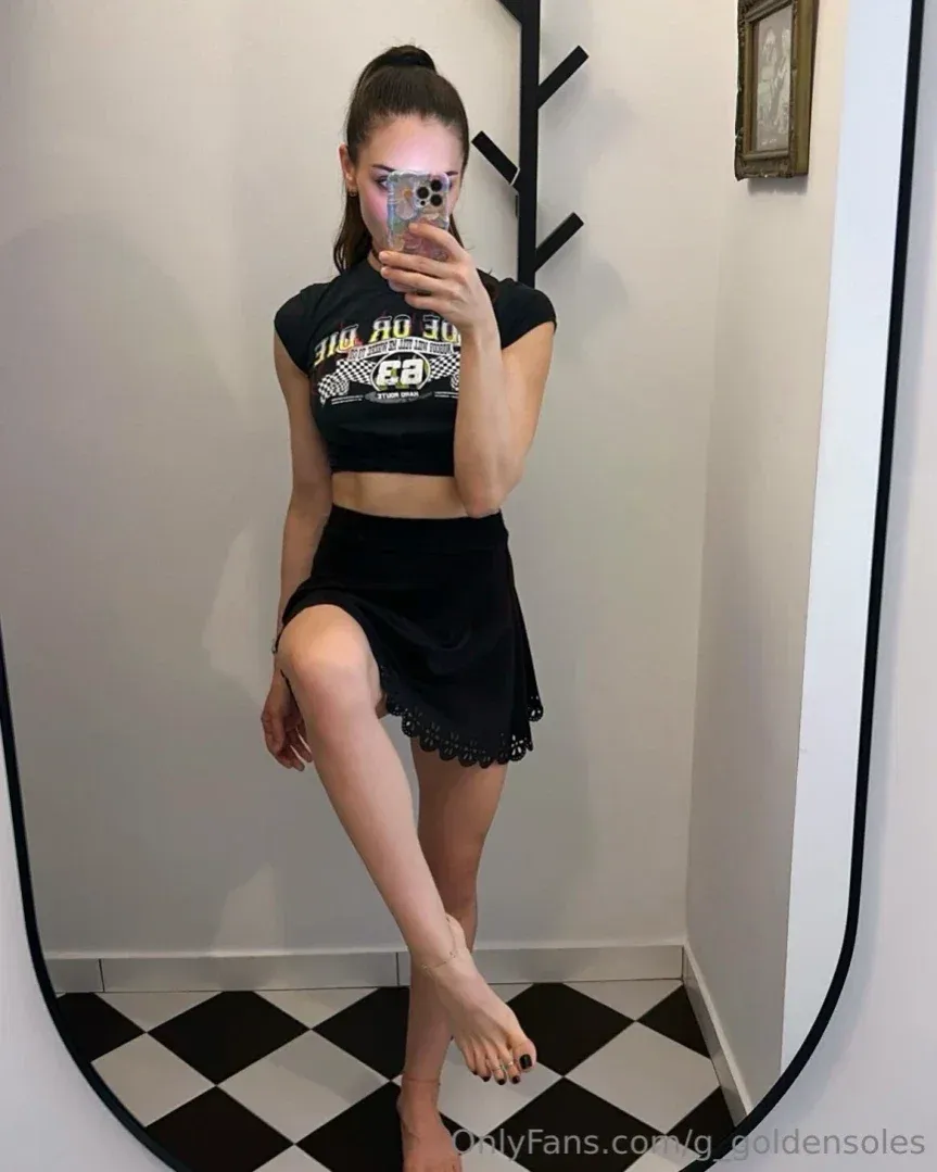 Gianna prend un selfie dans le miroir avec un crop top et une jupe noirs, debout sur un sol à damier.
