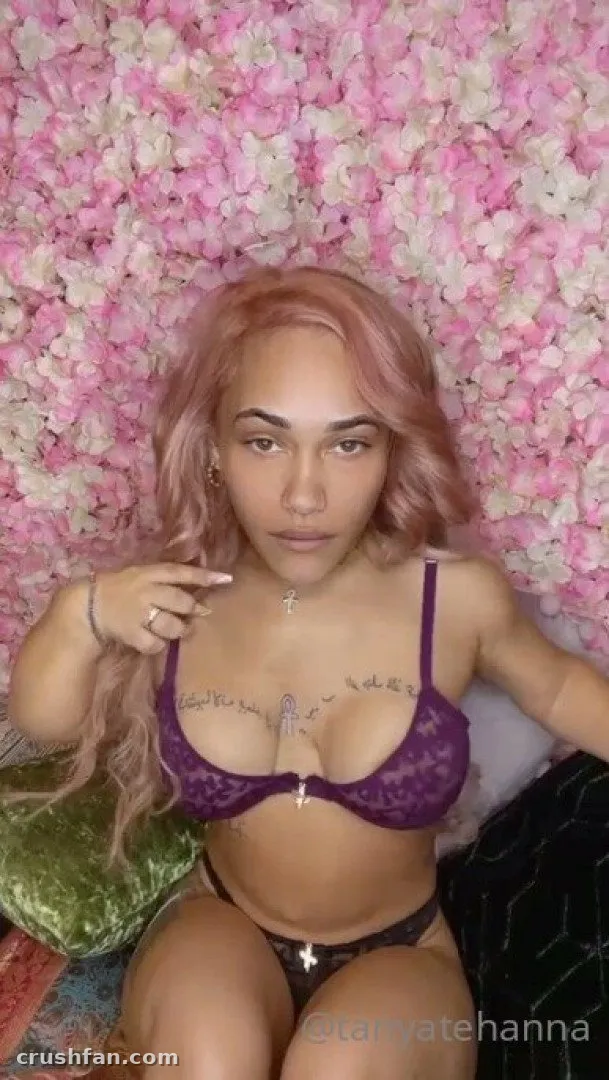 Tanya en lingerie en dentelle violette assise devant un mur de fleurs roses et blanches