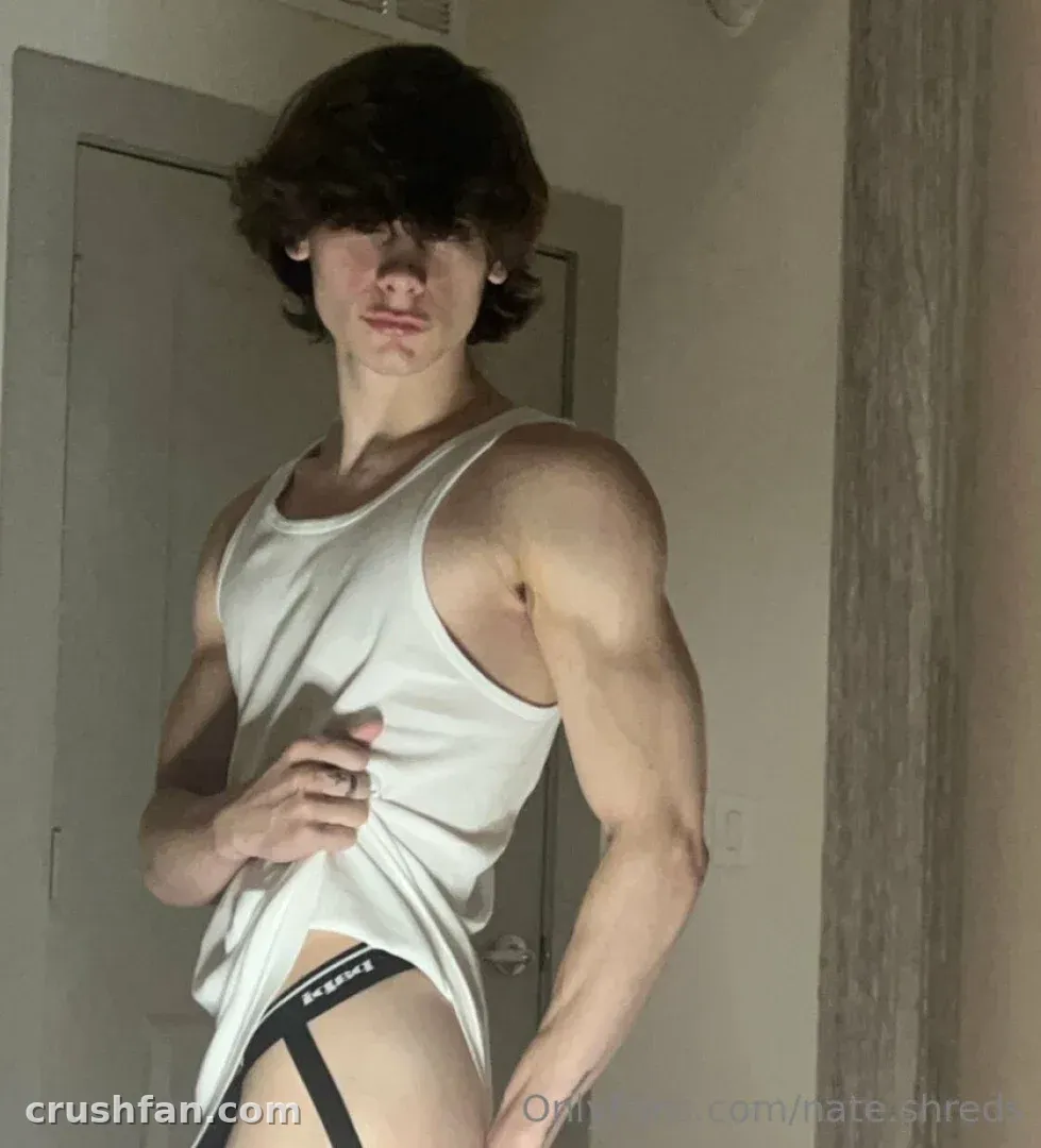 NATHAN in a white tank top and dark briefs standing indoors by a grey door