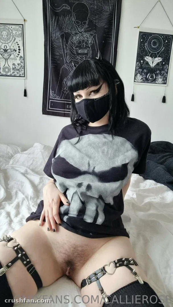 Callie Rose com camiseta preta de caveira e máscara facial reclinada em uma cama com tapeçarias góticas na parede
