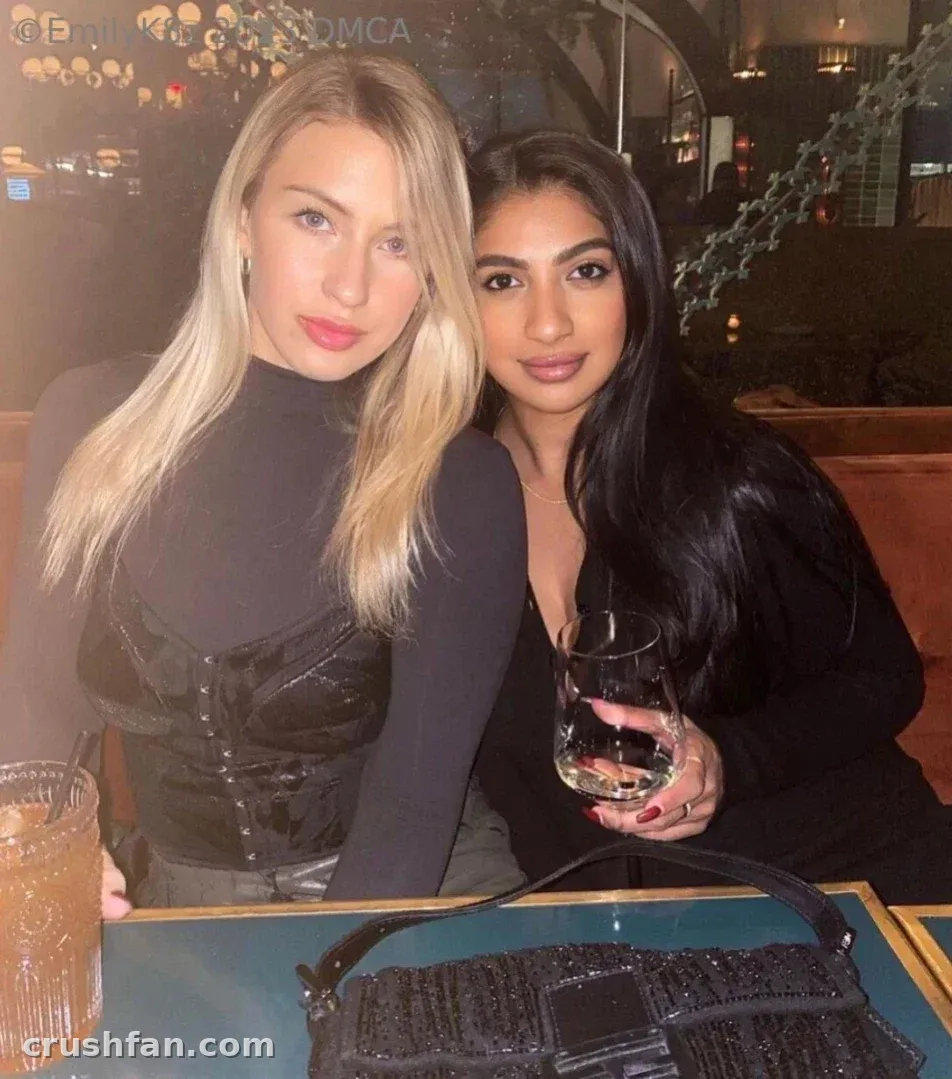 Emma K sitzt an einem Tisch in einer schwach beleuchteten Lounge neben einer anderen Frau, beide blicken zur Kamera.