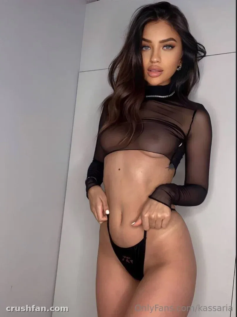 Kessi vestindo um top preto transparente e uma tanga combinando contra uma parede clara