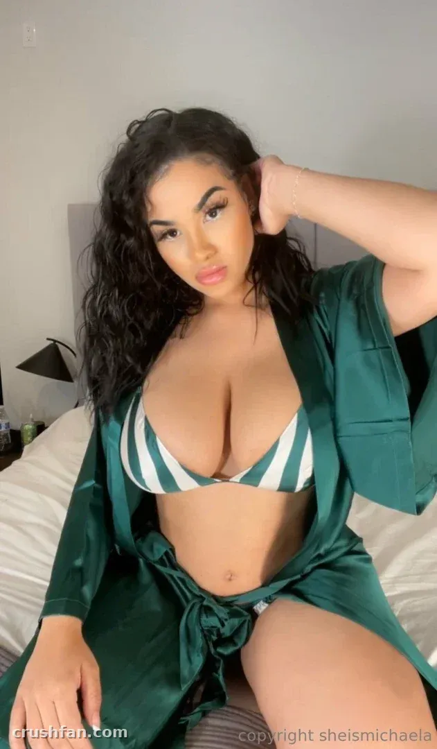 Michaela dans une robe de satin vert et un haut de bikini rayé assise sur un lit à l'intérieur