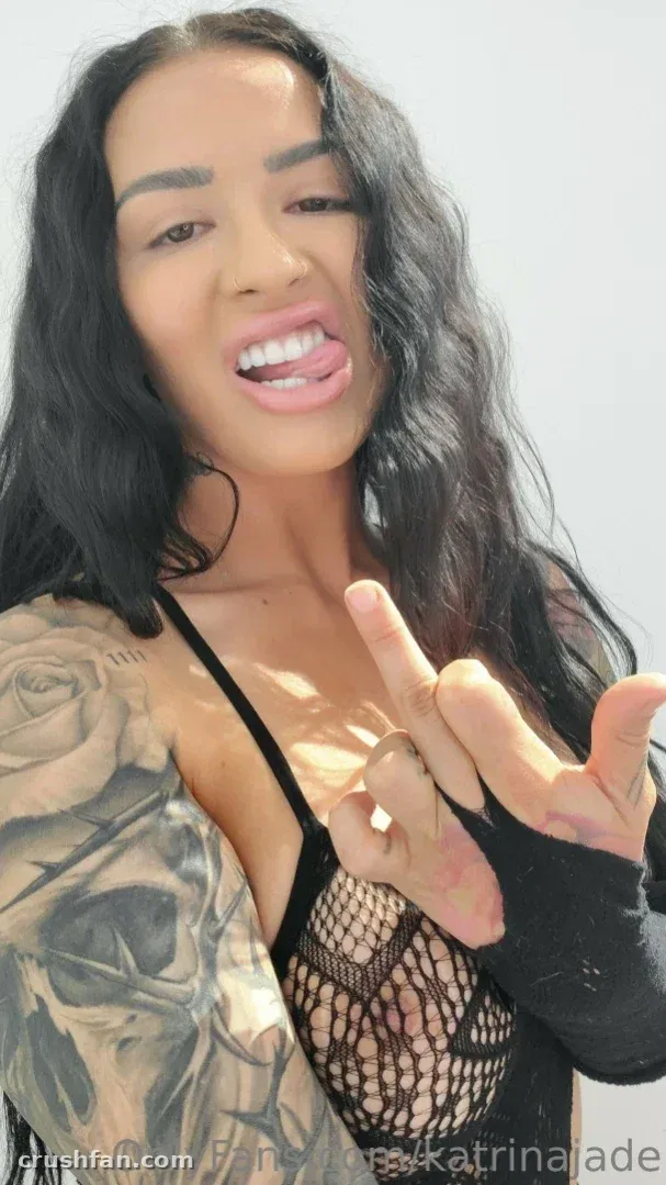 Katrina Jade in a black fishnet top making a hand gesture against a light background