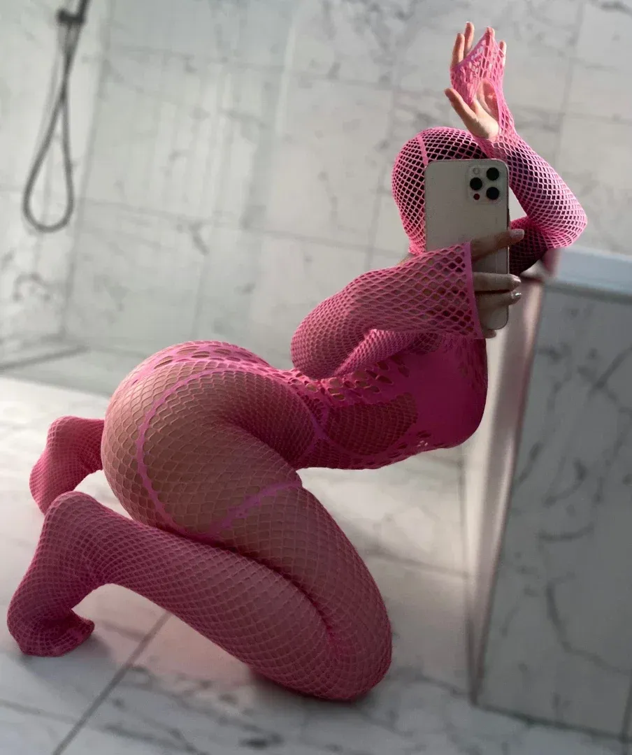 Ginasavagex con un bodystocking de red rosa arrodillada en un suelo de mármol tomándose un selfie en el espejo