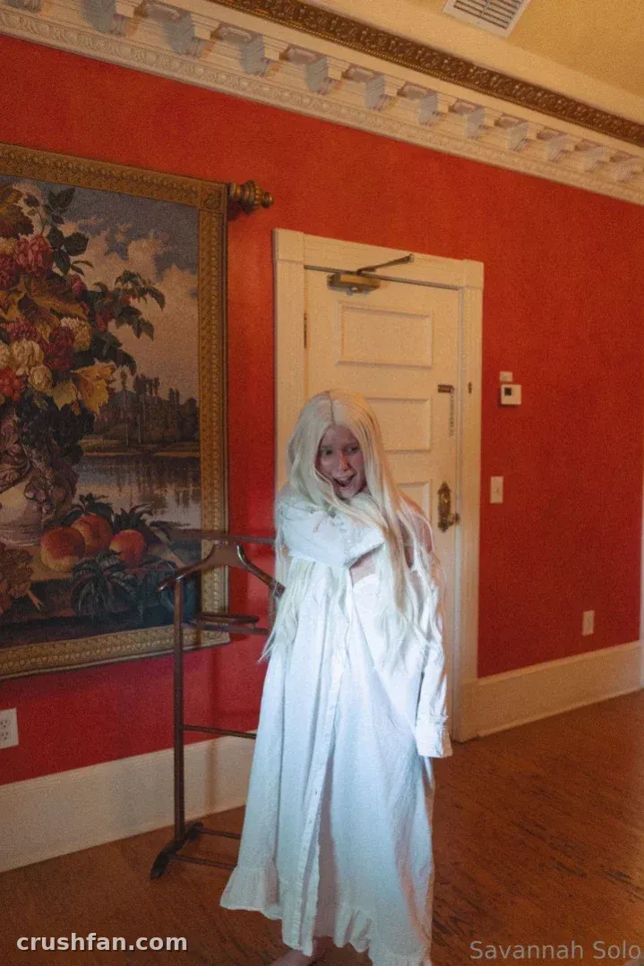 Savannah Solo em um vestido branco, de pé em uma sala vermelha com uma pintura emoldurada e uma porta branca.