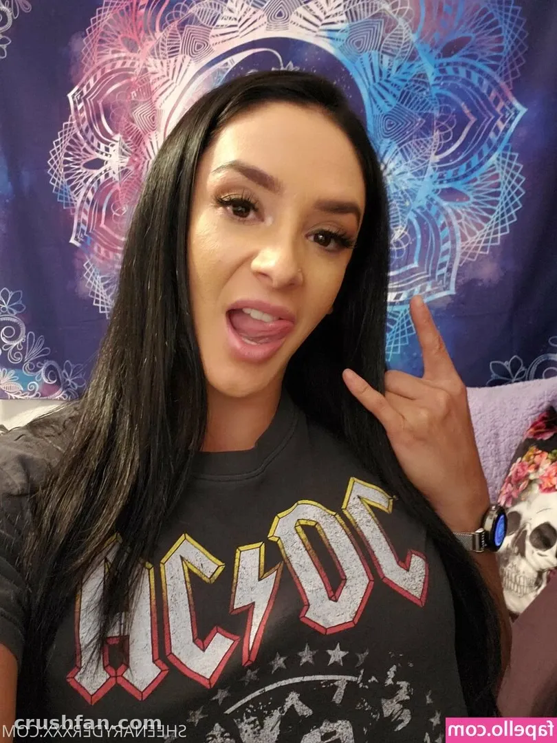 Sheena Ryder com uma camiseta gráfica escura fazendo um gesto de rock em frente a uma tapeçaria mandala azul