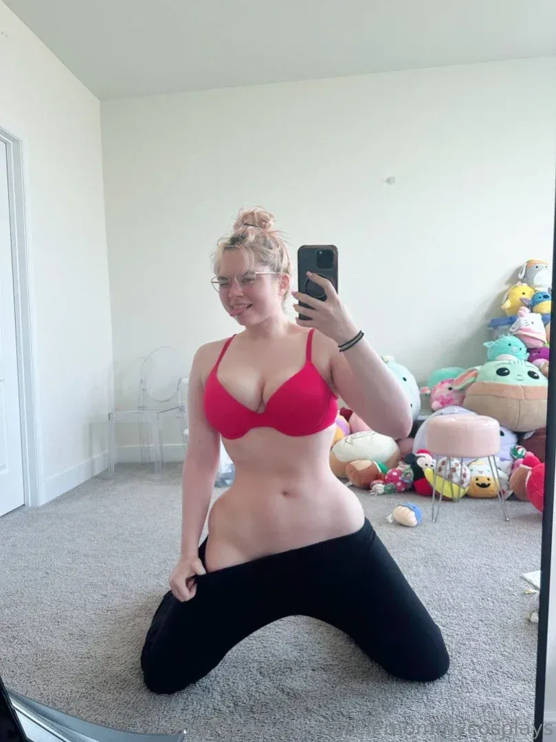 Linnea kneels on a carpet in a red bra and black leggings, taking a mirror selfie in a bright room.