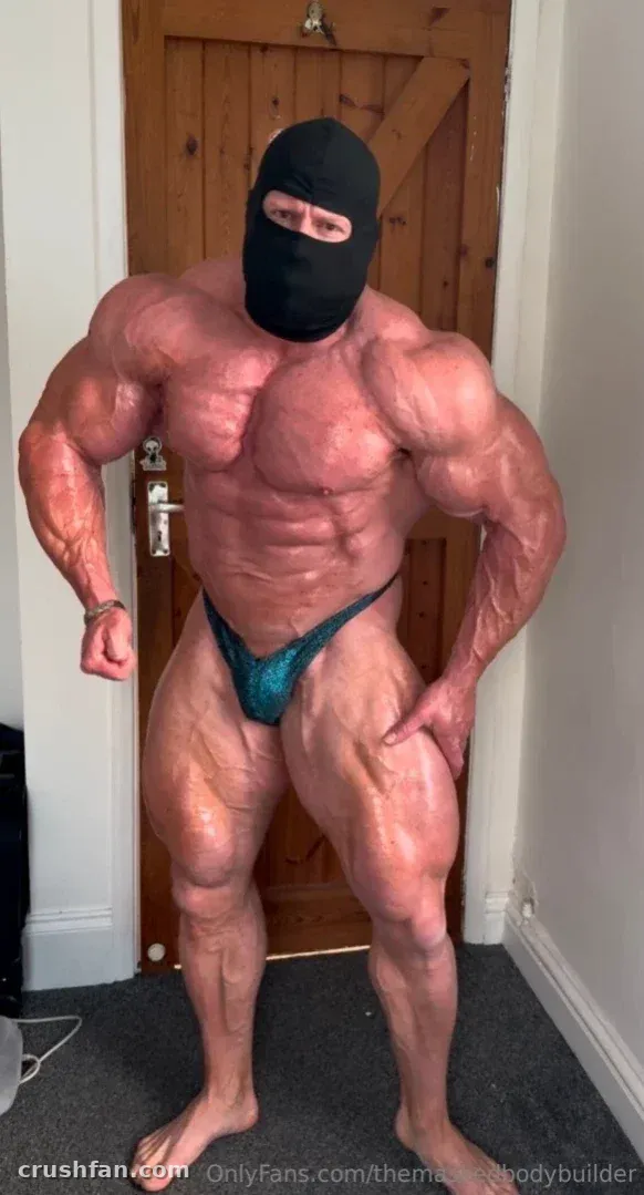 Muscular masked man in a teal posing pouch standing in a hallway.