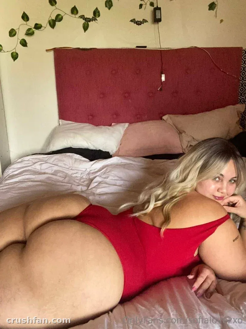 Sofia in a red bodysuit lying on a bed with white sheets and a red headboard