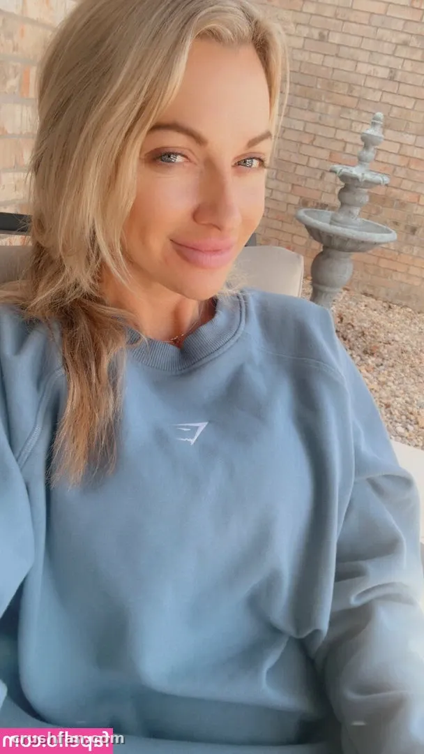 Blonde woman in a light blue sweatshirt posing outdoors by a brick wall and fountain