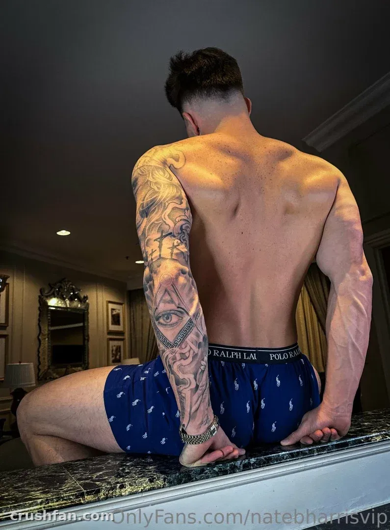 Nate assis torse nu sur une surface en marbre, montrant un bras tatoué et un boxer bleu marine.