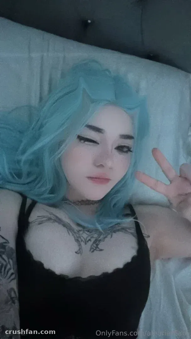 Criadora com peruca azul ondulada e top preto apoiada sobre roupa de cama branca, fazendo um sinal de paz