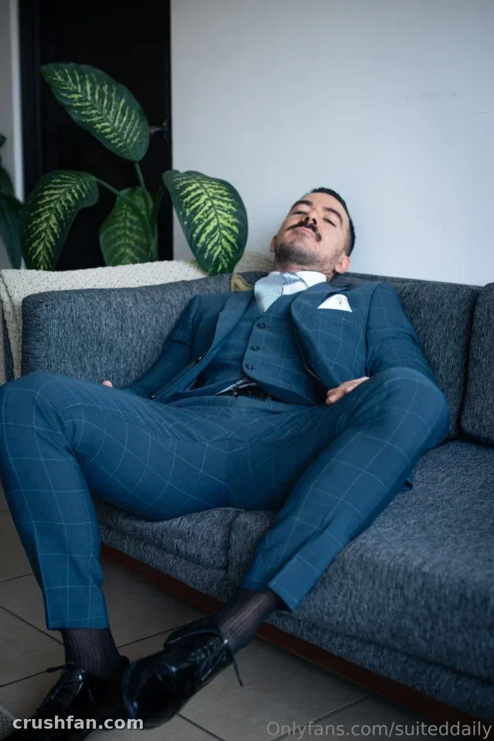 Man in a blue checked three-piece suit reclining on a grey sofa beside a leafy plant