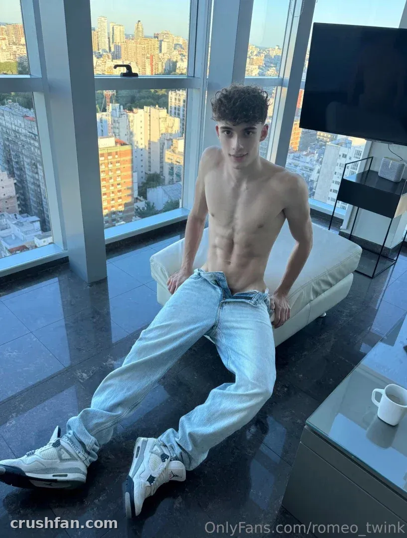 Romeo seated shirtless on a sofa in a high-rise room with city views through large windows.