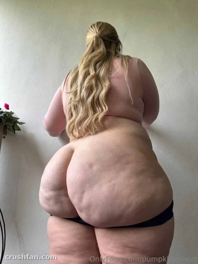 Pumpkin Cakezz seen from behind in a pink top and dark underwear beside a small plant.