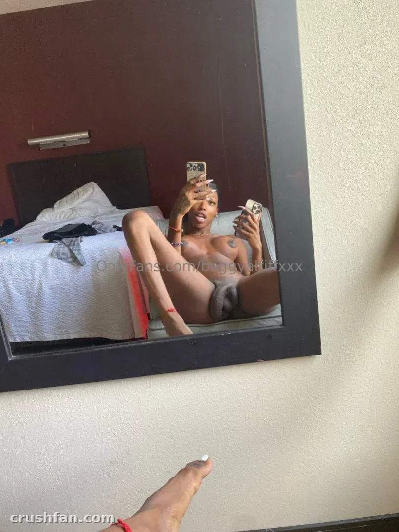 mAmi-Tiff prenant un selfie miroir tout en étant assise sur un lit d'hôtel dans une chambre avec un mur rouge.