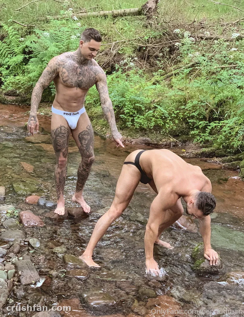Ben Dudman em pé em um riacho raso na floresta ao lado de um homem de roupa de banho preta inclinado sobre pedras molhadas