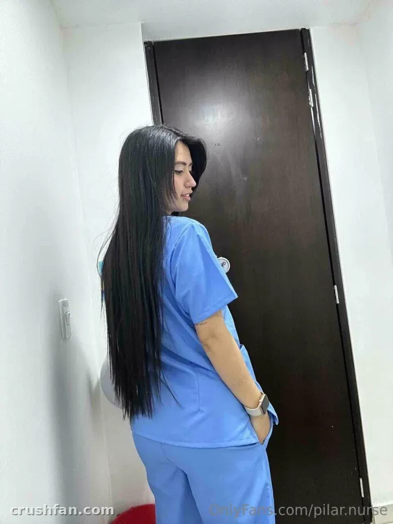 La enfermera Pilar con uniforme azul de pie junto a una puerta de madera oscura en una habitación interior luminosa