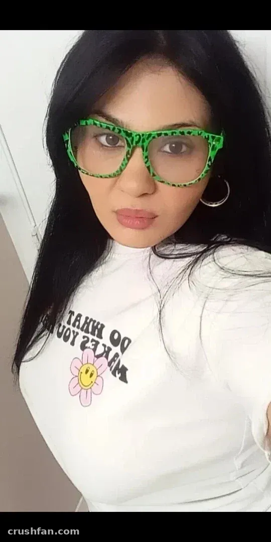 Beautybygen avec des lunettes vertes à motifs et un haut graphique blanc, prenant un selfie à l'intérieur.