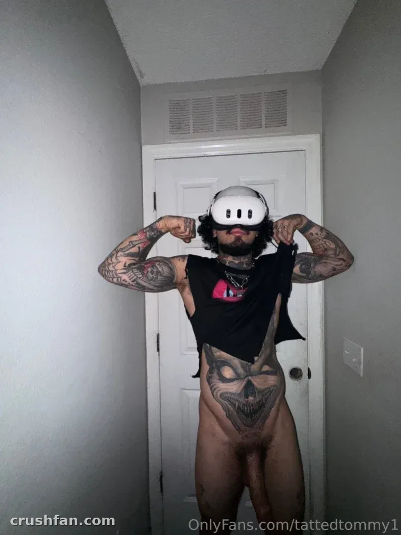 Tommy con visor de realidad virtual y top negro corto flexionando sus brazos tatuados en un pasillo
