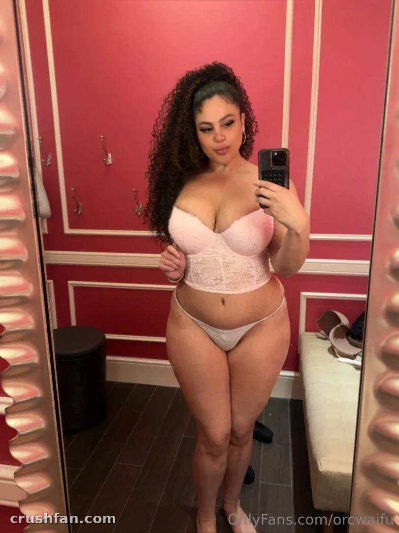 Gabby Z de lingerie de renda branca tirando uma selfie no espelho em uma sala com painéis vermelhos