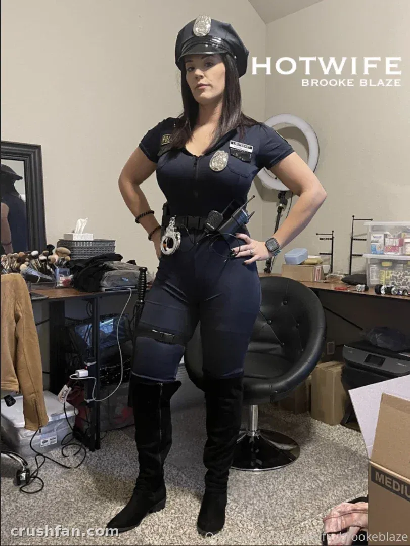 Hotwife Brooke Blaze poses in a navy police costume with a cap and tall black boots indoors.
