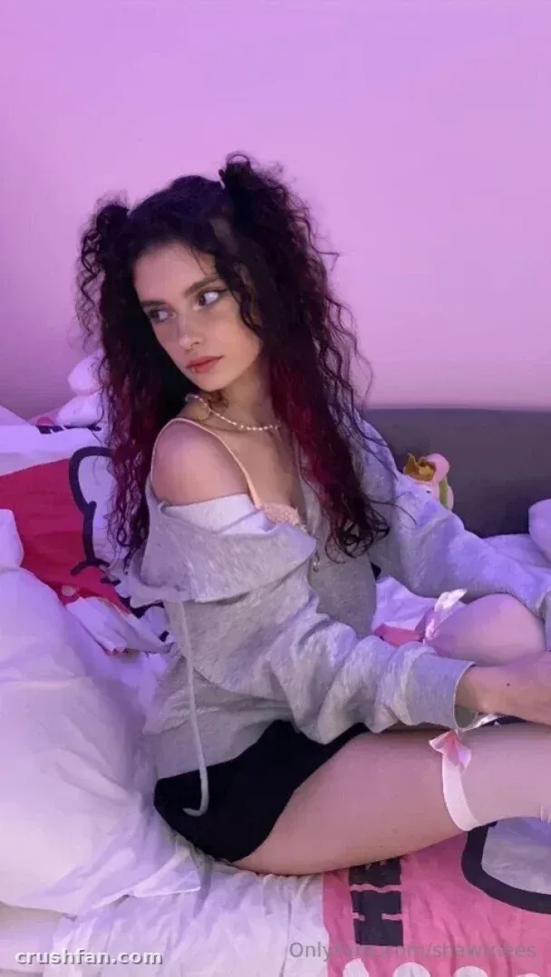 Nestix sitting on a bed in a lilac-lit room wearing a grey off-shoulder top and dark shorts