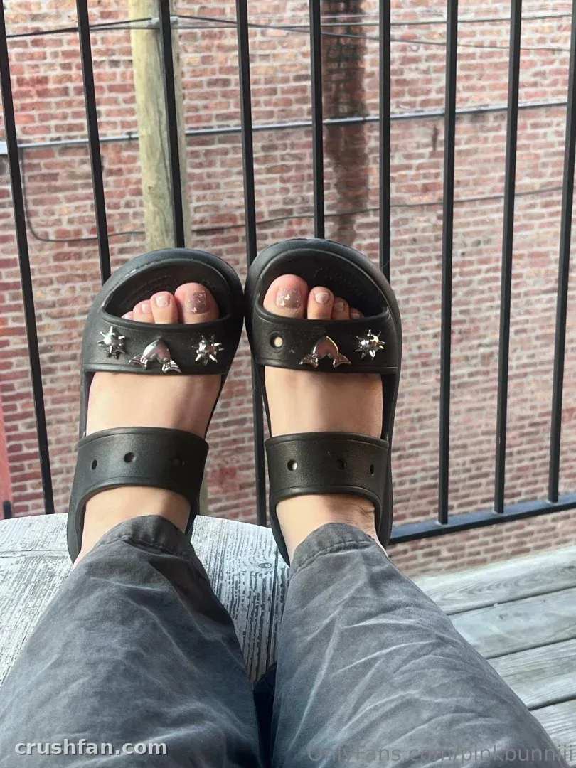 Nahaufnahme von schwarzen nietenbesetzten Sandalen und grauer Hose vor einer Backsteinmauer und einem Geländer.