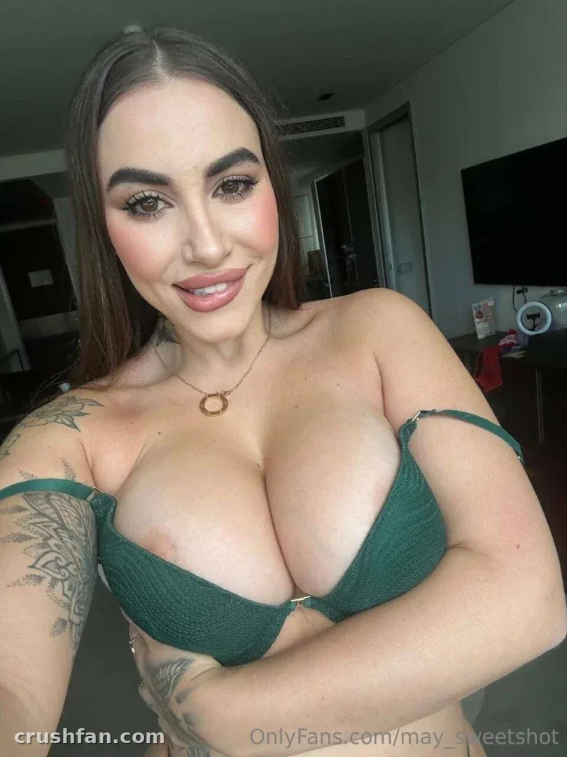 Creadora con un top verde sonriendo en una selfie en interiores con tatuajes visibles.