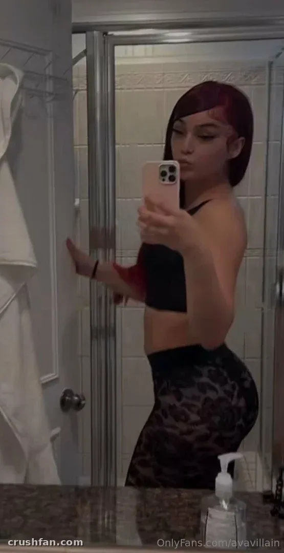 Miss Throat Goat prenant un selfie dans le miroir dans une salle de bain, portant un top noir et un legging à motifs.