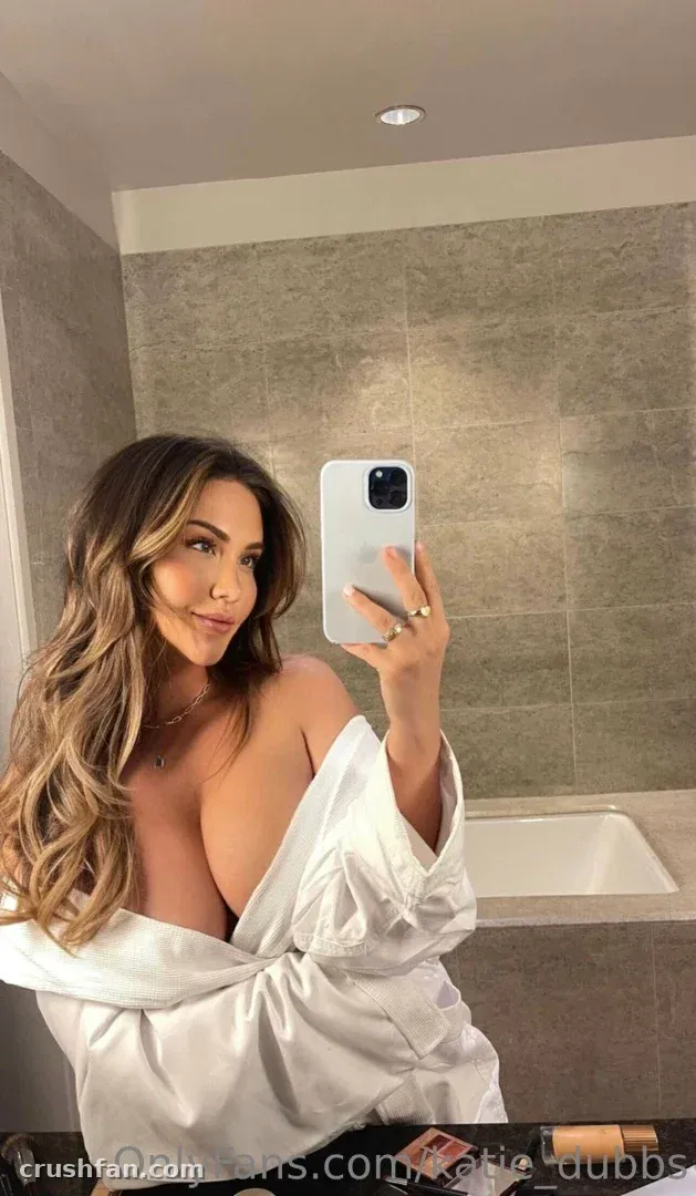 Katie en peignoir blanc prenant un selfie dans le miroir d'une salle de bain carrelée.