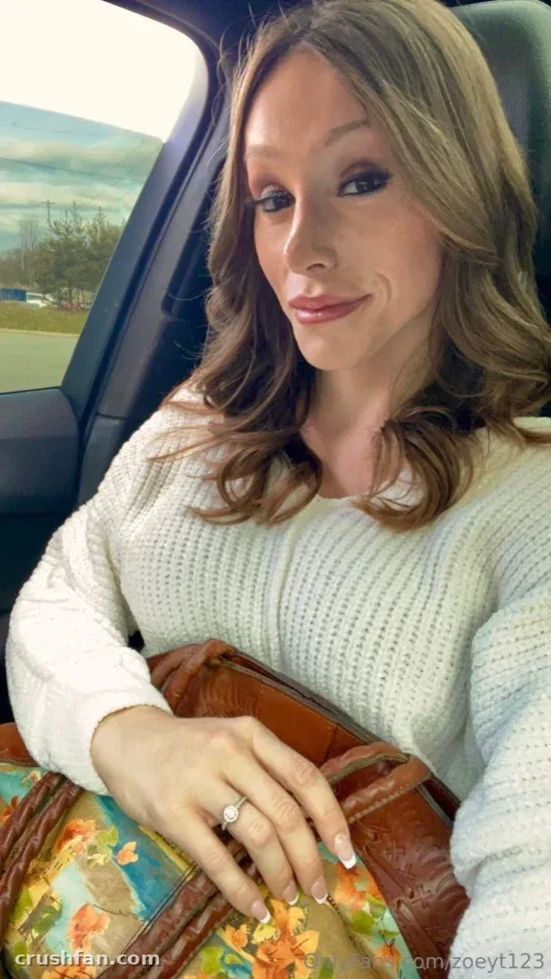 Zoey Taylor com um suéter de tricô cor creme sentada em um carro com uma bolsa de couro marrom no colo.