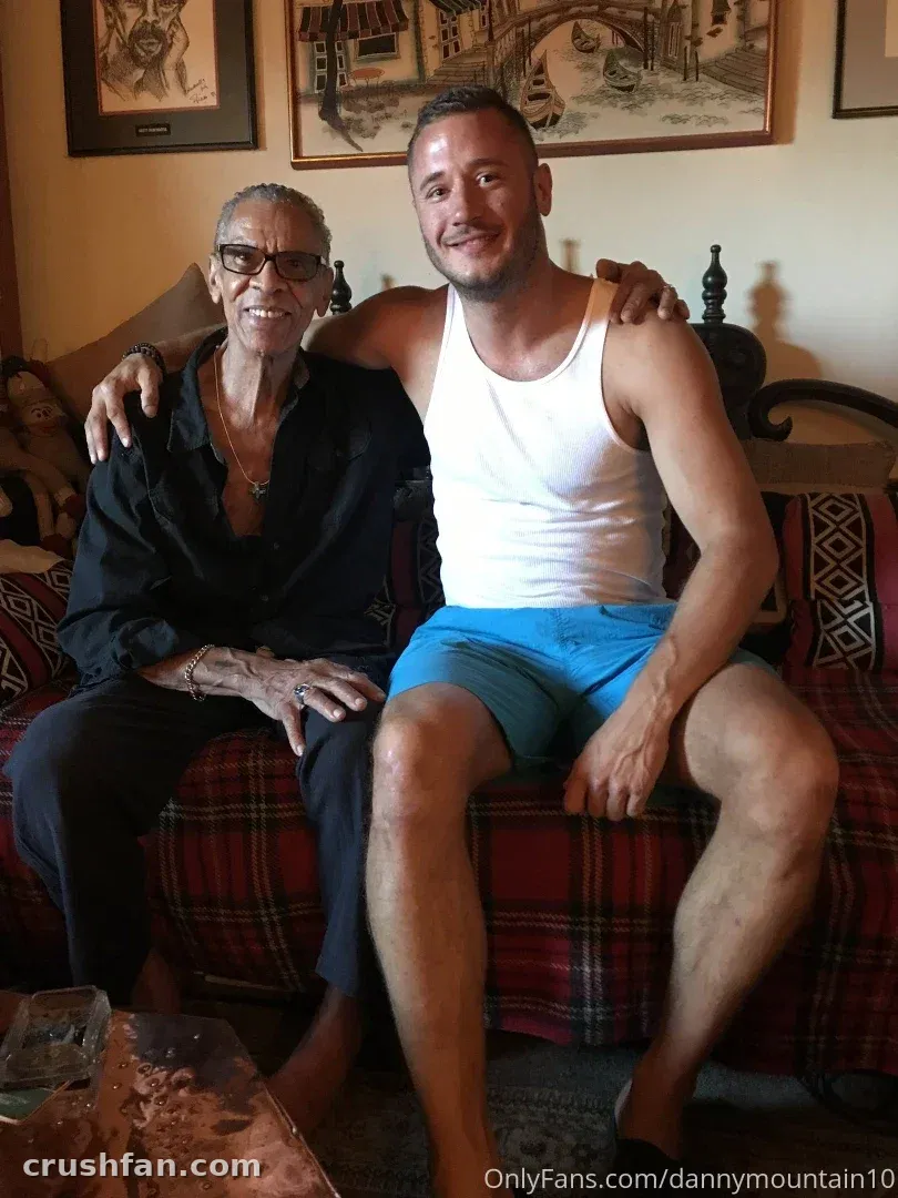 Danny Mountain con una camiseta sin mangas blanca y pantalones cortos azules sentado en un sofá a cuadros en el interior.