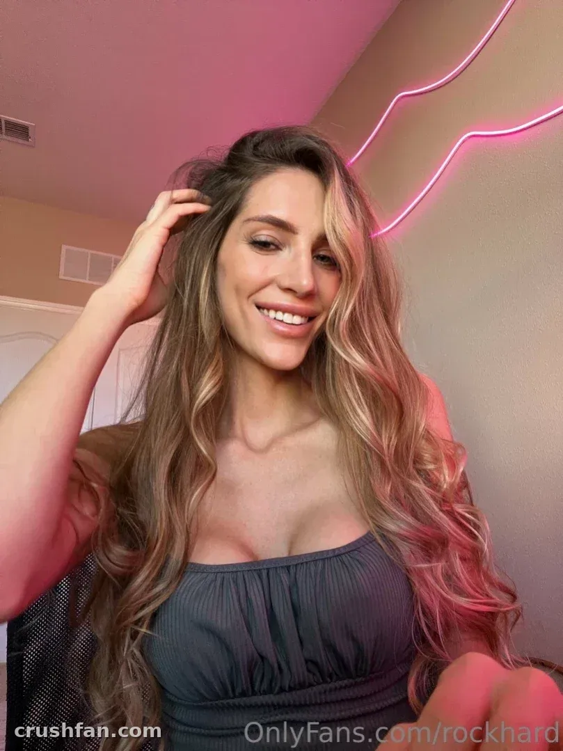 Holly Roxx souriant à l'intérieur sous une lumière néon rose, portant un haut sombre