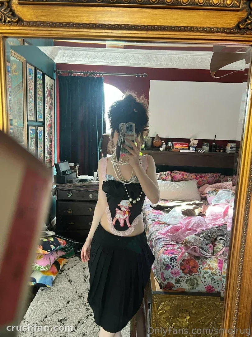 Creator in einem schwarzen Top und Rock, die in einem Schlafzimmer mit rosa Bettwäsche ein Spiegel-Selfie macht.