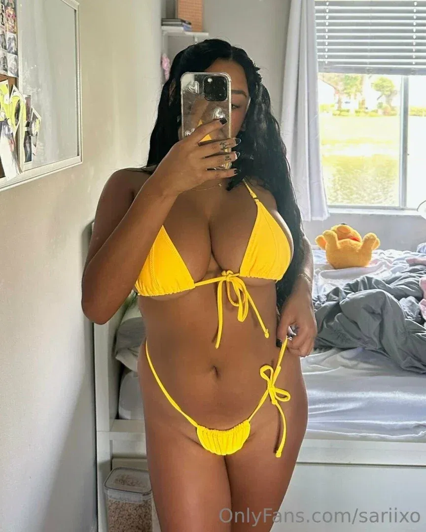Sari tomándose una selfie frente al espejo con un bikini de tiras amarillo dentro de un dormitorio.