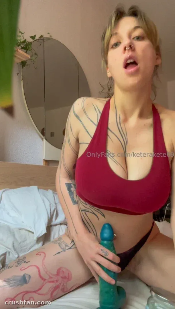 La créatrice KAT sur un lit dans un haut à dos nu bordeaux avec des tatouages visibles, en intérieur