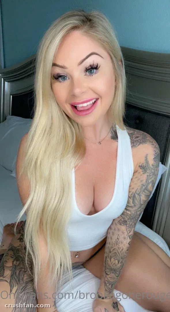 Brooklyn com um top cropped branco sentada em uma cama com uma parede azul atrás dela.