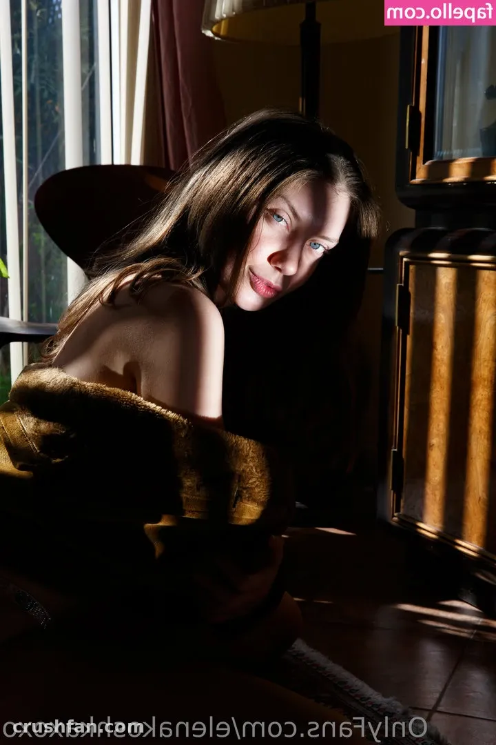 Elena Koshka seated by a window in soft daylight, wrapped in a golden-brown fabric