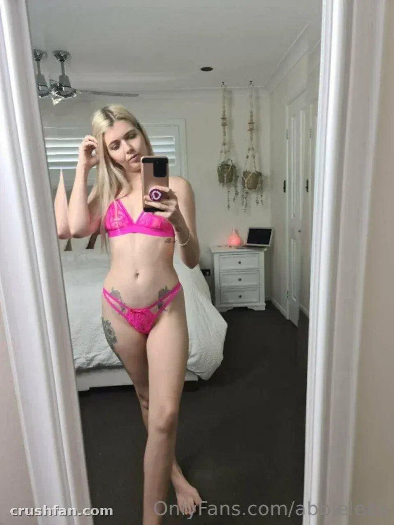 Abbie en lingerie en dentelle rose prenant un selfie miroir dans une chambre.