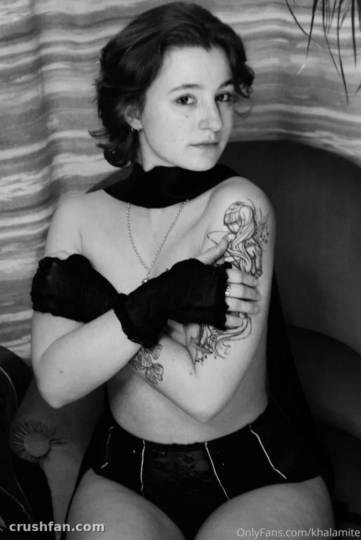 Black and white portrait of Khalamité seated with arms crossed, wearing dark gloves and a scarf.