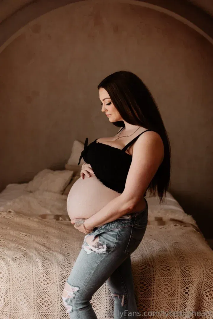 Lyla in a black top and ripped jeans standing beside a bed in a softly lit room
