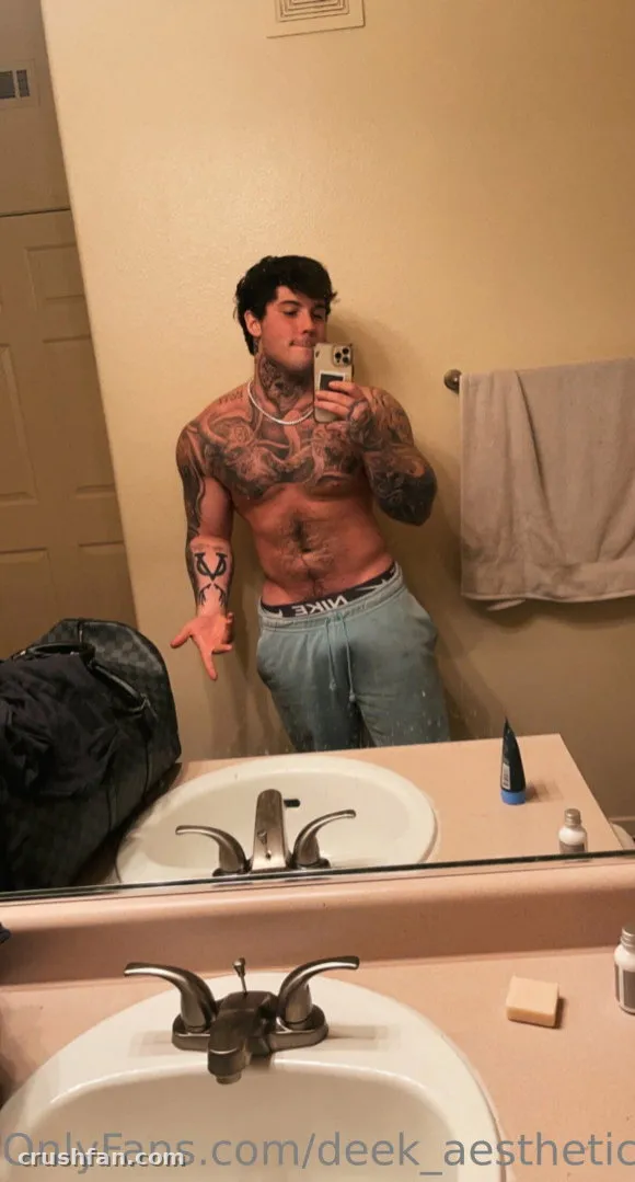 DEEK AESTHETIC macht ein Spiegel-Selfie in einem Badezimmer und zeigt Tattoos und Jogginghose.