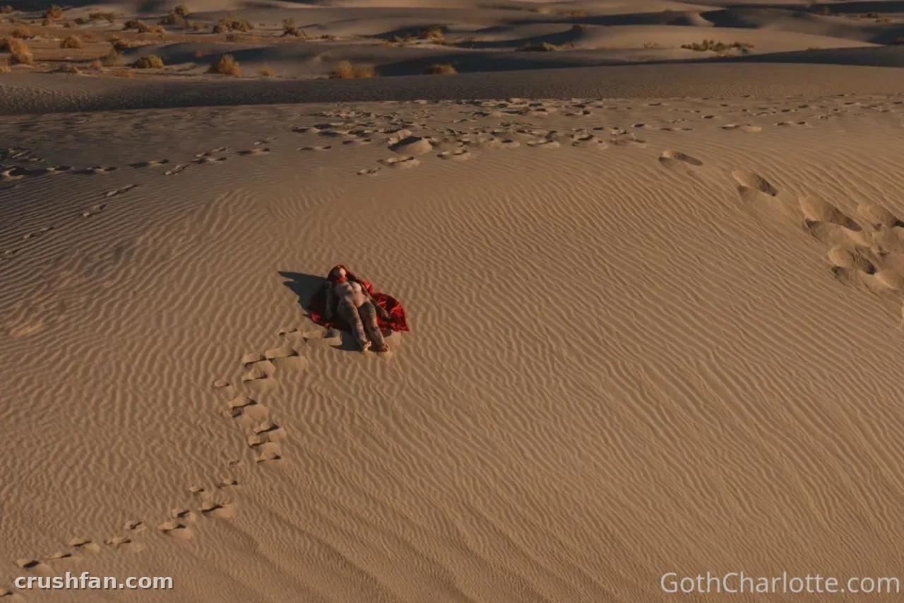 Goth Charlotte sentada sobre areia ondulada do deserto com um tecido vermelho ao seu lado ao pôr do sol