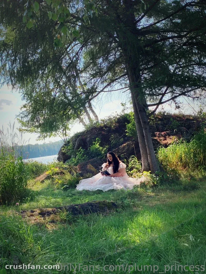 Plump Princess in a pale pink gown seated on grass under a tree by a lake