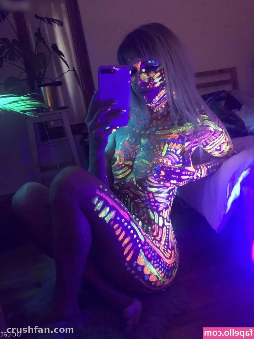 Jana tira uma selfie no espelho enquanto seu corpo está coberto de pintura corporal neon brilhante sob luz negra.