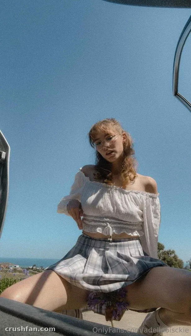 Adella in a white off-shoulder top and plaid skirt outdoors under a clear blue sky